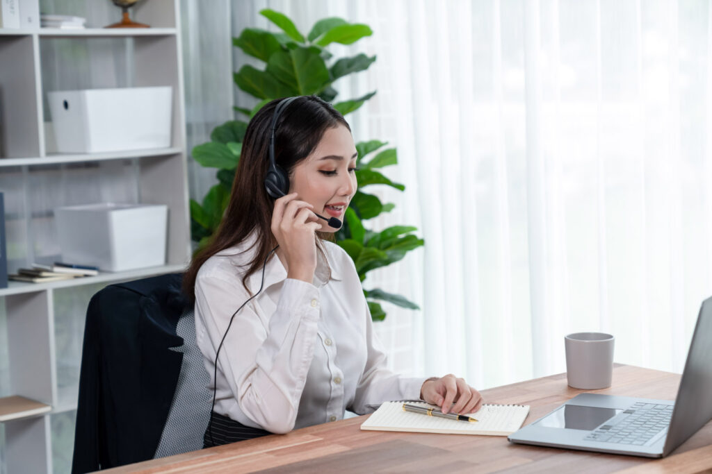 Asian doing outbound call center services with headset and microphone working on her laptop. Female operator provide exceptional customer service. Supportive call center agent helping customer on inquiry. Back Office Administration