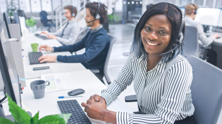 Beautiful Young Female Customer Service Operator Smiling for a Portrait at a Busy Modern Call Center with Diverse Multicultural Team of Office Specialists Wearing Headsets and Taking Calls.
